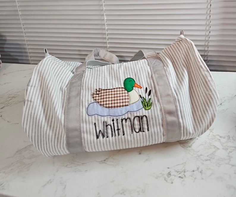 May include: A striped duffel bag with a duck applique. The duck has a green head, brown body, and orange beak. The bag has gray trim and handles, and the name "Whitman" is embroidered below the duck.