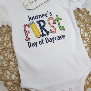 K&ouml;nnte beinhalten: Wei&szlig;er Strampler mit einem bunten gestickten Design, auf dem "Journee's First Day of Daycare" steht.