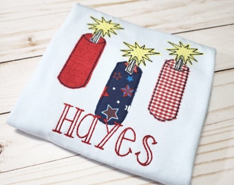 May include: A light blue t-shirt with three red, white, and blue firecracker designs. The name "Hayes" is embroidered in red on the shirt.