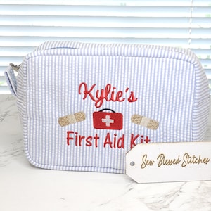 May include: A blue and white striped first aid kit bag embroidered with "Kylie's" in red script. The bag features a red first aid kit graphic with a white cross and two embroidered bandages. The words "First Aid Kit" are also embroidered in red.