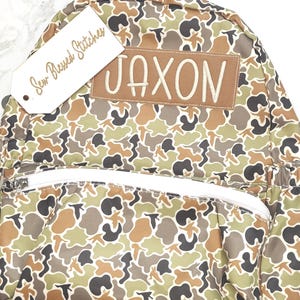 Puede incluir: Una mochila con estampado de camuflaje con cremallera y correas blancas. La mochila presenta un parche marr&oacute;n con el nombre "JAXON" bordado en blanco. Una etiqueta que dice "Sew Blessed Stitches" est&aacute; adjunta.