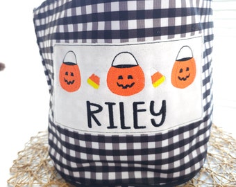 May include: A black and white gingham fabric Halloween bucket with a white patch featuring three orange candy corn buckets and the name "RILEY" embroidered in black. The bucket is sitting on a woven mat.
