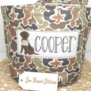 May include: A camouflage-patterned tote bag featuring the name "COOPER" embroidered in black script on a white patch. A brown dog with an Easter basket is embroidered below the name. The tag reads "Sew Blessed Stitches".