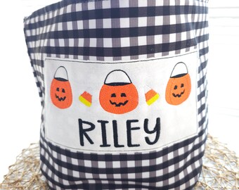 May include: Black and white gingham fabric Halloween treat bag with three embroidered orange pumpkins and candy corn. The bag has the name "Riley" embroidered on the front.