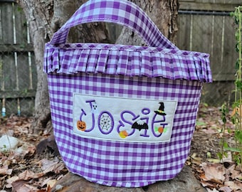 May include: Purple and white gingham trick-or-treat bag with a ruffled trim and the name "Josie" embroidered on the front. The embroidery includes a ghost, spider web, pumpkin, bat, and witch's hat.