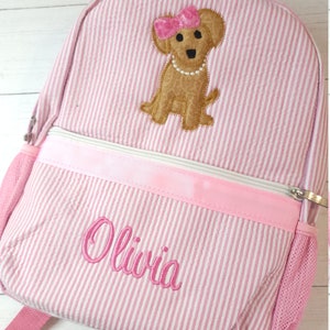 May include: Pink and white striped backpack with a brown dog embroidered on the front. The dog is wearing a pink bow and a pearl necklace. The backpack has a pink zipper and a mesh pocket on the side. The name "Olivia" is embroidered on the front of the backpack.