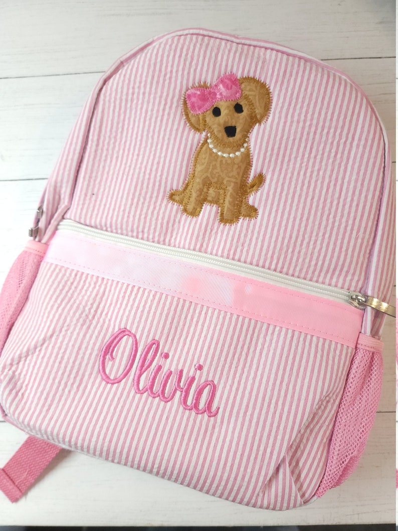 May include: Pink and white striped backpack with a brown dog embroidered on the front. The dog is wearing a pink bow and a pearl necklace. The backpack has a pink zipper and a mesh pocket on the side. The name "Olivia" is embroidered on the front of the backpack.
