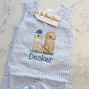Pu&ograve; includere: Una tutina in seersucker a righe azzurre e bianche con un ricamo di due cani, uno che indossa un berretto da baseball blu e l'altro con una mazza e una palla. Il nome Decker &egrave; ricamato in blu. Un'etichetta riporta la scritta Sew Blessed Stitches.