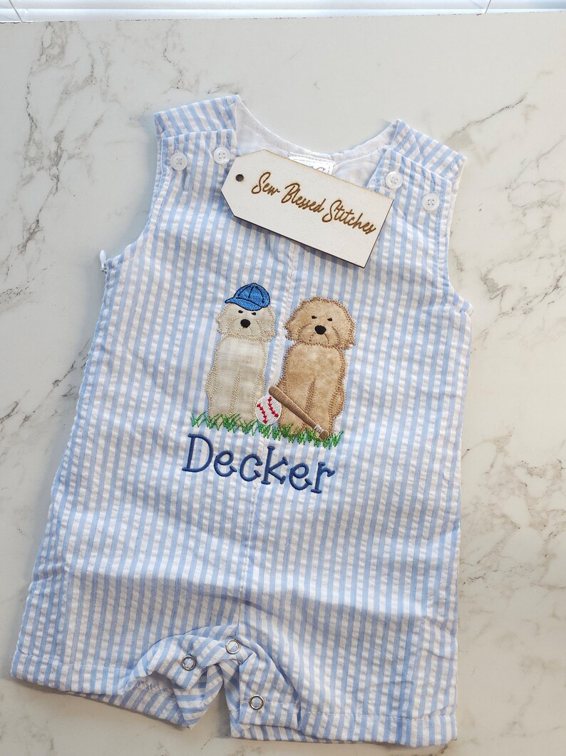 Pu&ograve; includere: Una tutina in seersucker a righe azzurre e bianche con un ricamo di due cani, uno che indossa un berretto da baseball blu e l'altro con una mazza e una palla. Il nome Decker &egrave; ricamato in blu. Un'etichetta riporta la scritta Sew Blessed Stitches.