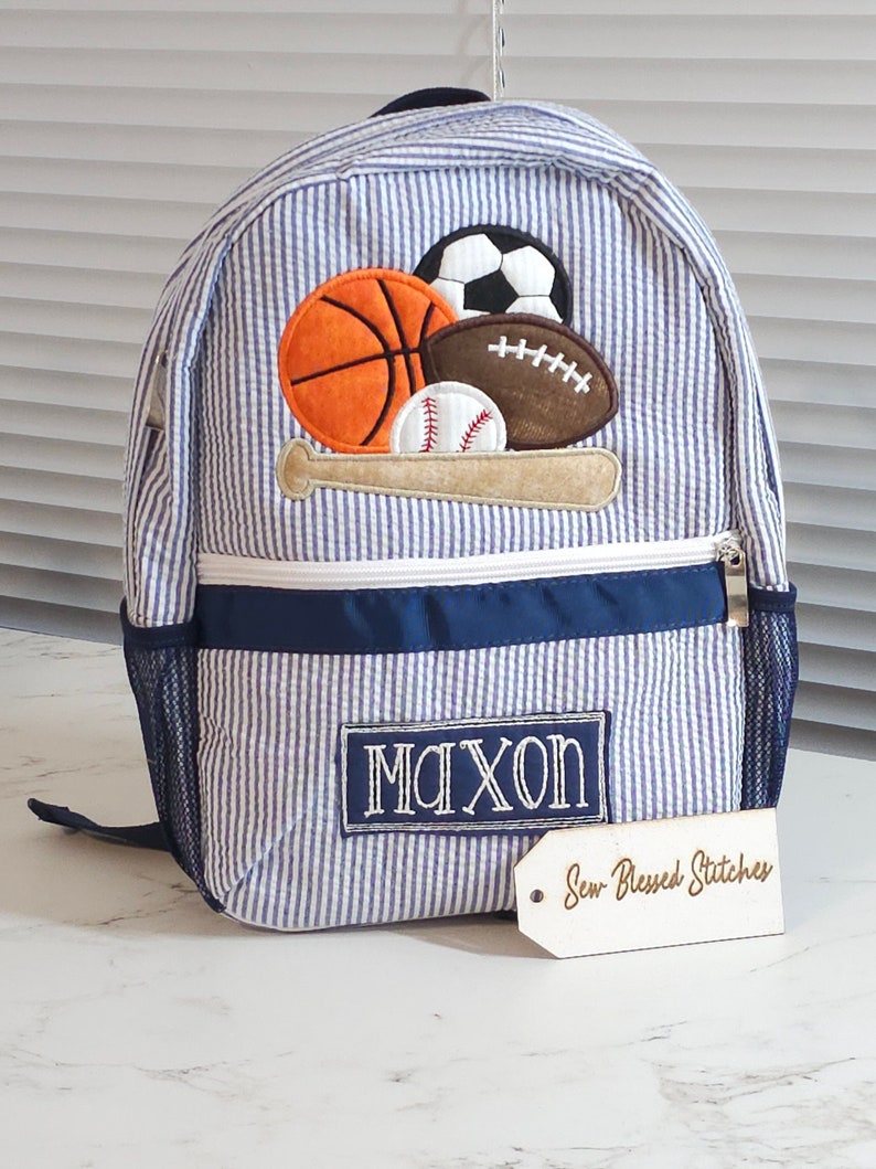 May include: Blue and white striped backpack with a navy blue trim. The backpack has a large embroidered patch with a baseball bat, baseball, basketball, football, and soccer ball. The backpack also has a navy blue pocket with the name "Maxon" embroidered on it. A white tag with the text "Sew Blessed Stitches" is attached to the backpack.
