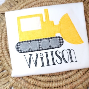 May include: White t-shirt featuring a yellow bulldozer design with black and white checkered tracks and the name "Willson" embroidered in black. The shirt is on a woven, tan surface.