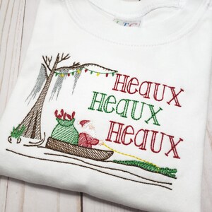May include: A white t-shirt featuring an embroidered design of Santa Claus in a pirogue boat pulled by an alligator. The scene includes a cypress tree with Spanish moss and Christmas lights. The text reads Heaux Heaux Heaux in red and green.