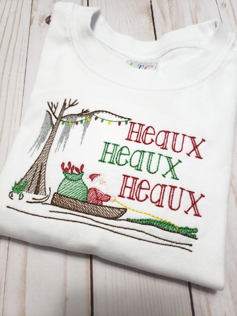 May include: A white t-shirt featuring an embroidered design of Santa Claus in a pirogue boat pulled by an alligator. The scene includes a cypress tree with Spanish moss and Christmas lights. The text reads Heaux Heaux Heaux in red and green.