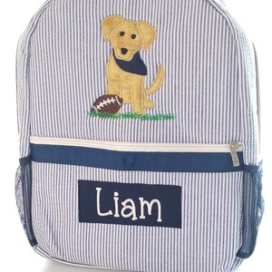 Peut inclure: Sac &agrave; dos ray&eacute; bleu et blanc avec un chien jaune et un ballon de football brod&eacute;s sur le devant. Le sac &agrave; dos a une poche zipp&eacute;e bleu marine et une poche lat&eacute;rale en filet. Le nom "Liam" est brod&eacute; sur la poche avant.