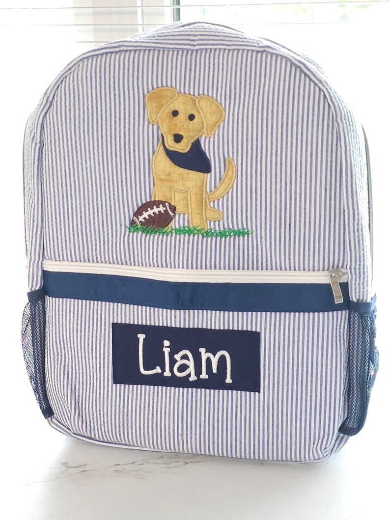 Peut inclure: Sac &agrave; dos ray&eacute; bleu et blanc avec un chien jaune et un ballon de football brod&eacute;s sur le devant. Le sac &agrave; dos a une poche zipp&eacute;e bleu marine et une poche lat&eacute;rale en filet. Le nom "Liam" est brod&eacute; sur la poche avant.