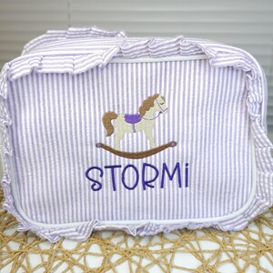 May include: A lavender and white striped storage bin with a rocking horse embroidery. The bin has a ruffled edge and the name "STORMI" embroidered in purple. The rocking horse is tan, with a purple saddle.