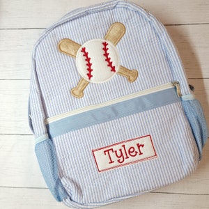 K&ouml;nnte beinhalten: Blau-wei&szlig; gestreifter Rucksack mit einem Baseball- und gekreuzten Fledermaus-Design. Der Rucksack hat eine blaue Netztasche an der Seite und ein rotes Namensschild mit der Aufschrift "Tyler".