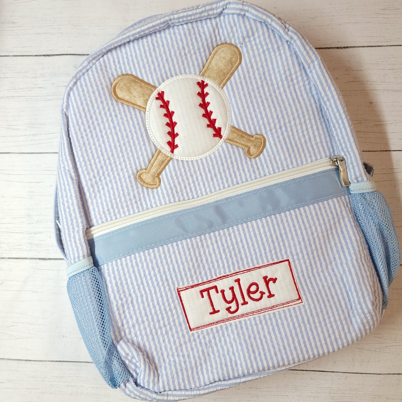 K&ouml;nnte beinhalten: Blau-wei&szlig; gestreifter Rucksack mit einem Baseball- und gekreuzten Fledermaus-Design. Der Rucksack hat eine blaue Netztasche an der Seite und ein rotes Namensschild mit der Aufschrift "Tyler".