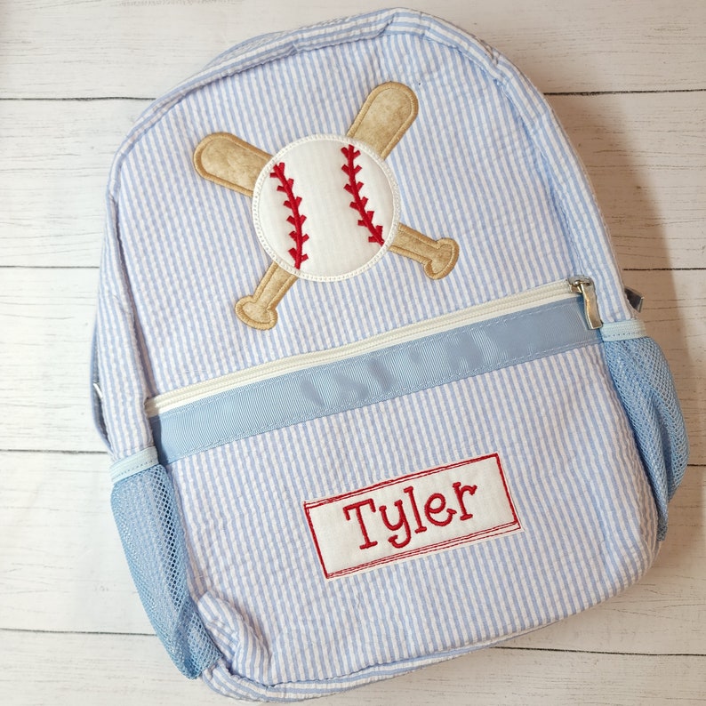 K&ouml;nnte beinhalten: Blau-wei&szlig; gestreifter Rucksack mit einem Baseball- und gekreuzten Fledermaus-Design. Der Rucksack hat eine blaue Netztasche und ein rotes Namensschild mit der Aufschrift "Tyler".