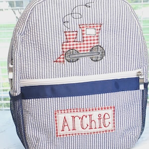 May include: A blue and white striped backpack with a red and white gingham train applique. The backpack has a blue zipper and a mesh pocket on the side. The name "Archie" is embroidered on the front of the backpack in red.