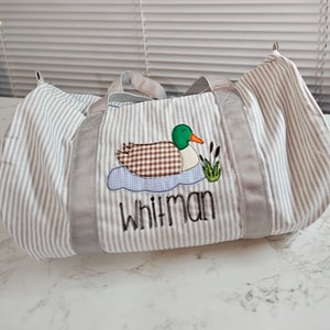 May include: A striped, gray and white duffel bag with a duck applique. The duck has a green head, brown body, and orange beak. The name "Whitman" is embroidered below the duck. The bag has gray handles and trim.