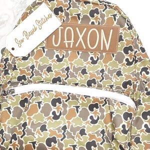Puede incluir: Una mochila con estampado de camuflaje con el nombre "JAXON" bordado en un parche marr&oacute;n. La mochila tiene una cremallera blanca y una etiqueta que dice "Sew Blessed Stitches". El patr&oacute;n incluye tonos de verde, marr&oacute;n y negro.