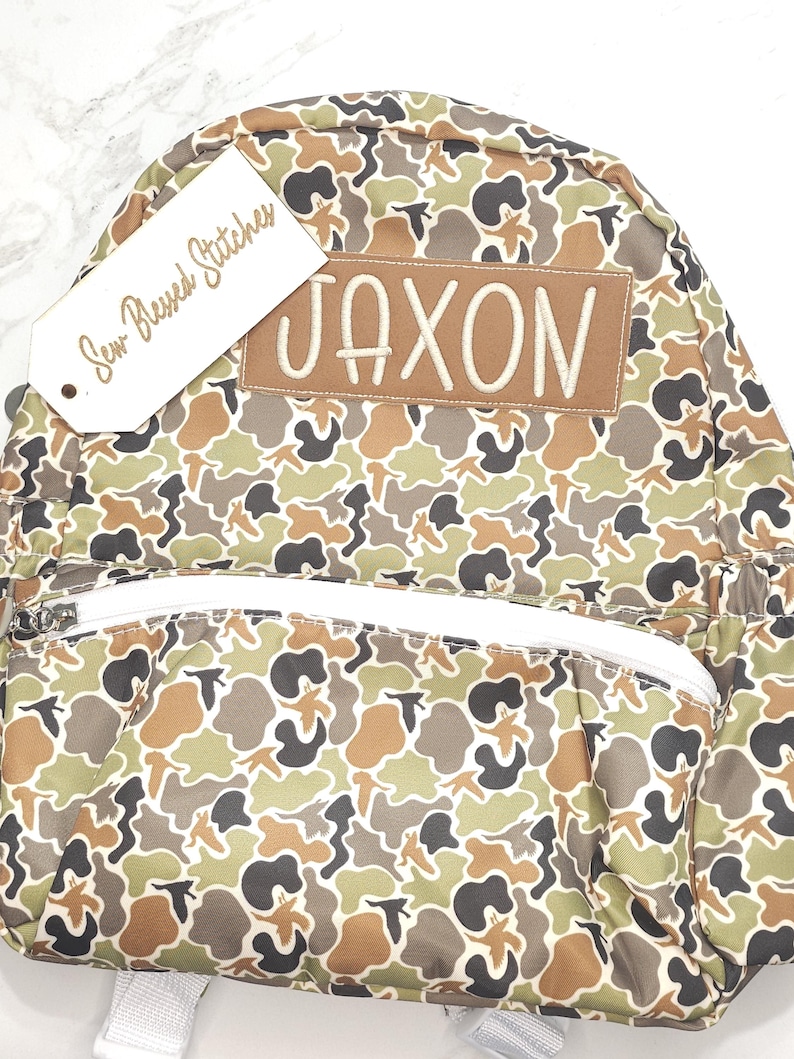 Puede incluir: Una mochila con estampado de camuflaje con el nombre "JAXON" bordado en un parche marr&oacute;n. La mochila tiene una cremallera blanca y una etiqueta que dice "Sew Blessed Stitches". El patr&oacute;n incluye tonos de verde, marr&oacute;n y negro.