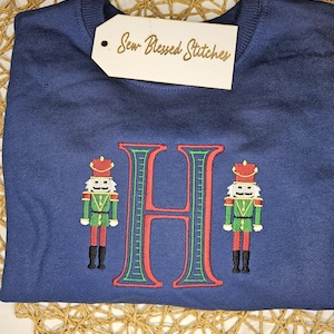 May include: A navy blue sweatshirt with an embroidered design. The design features a large red letter "H" with green accents, flanked by two embroidered nutcracker figures in red, green, and gold. A tag that says "Sew Blessed Stitches" is attached.