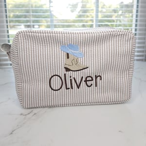 May include: A rectangular, seersucker-striped cosmetic bag with a brown and white pattern. The bag features an embroidered design of a cowboy boot and hat in blue and brown, with the name "Oliver" embroidered below.