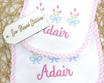 May include: White baby bibs with pink gingham trim and embroidered floral designs. The name "Adair" is embroidered in pink script. The tag reads "Sew Blessed Stitches".