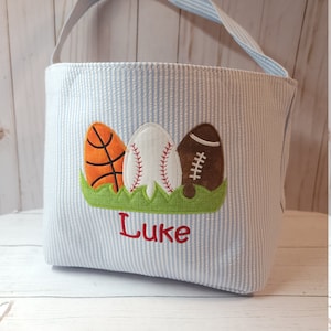 May include: A blue and white striped fabric tote bag with a handle. The bag has an embroidered design of three sports balls: a basketball, a baseball, and a football. The name "Luke" is embroidered below the balls.
