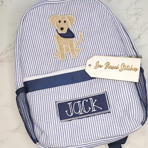 May include: A blue and white striped backpack with a dog applique. The backpack has a navy blue trim, a side mesh pocket, and the name "JACK" embroidered on a patch. A wooden tag reads "Sew Blessed Stitches".
