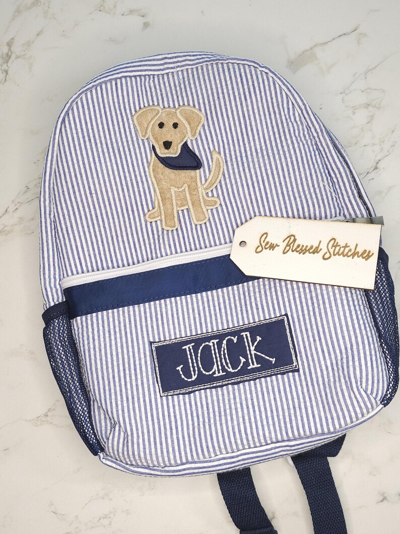 May include: A blue and white striped backpack with a dog applique. The backpack has a navy blue trim, a side mesh pocket, and the name "JACK" embroidered on a patch. A wooden tag reads "Sew Blessed Stitches".