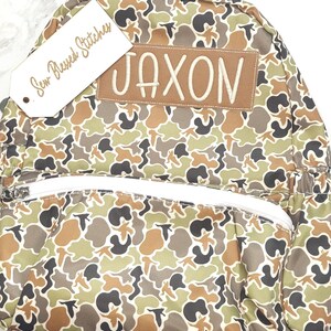 Puede incluir: Una mochila con estampado de camuflaje con el nombre "JAXON" bordado en un parche marr&oacute;n. La mochila tiene una cremallera y correas blancas, y una etiqueta que dice "Sew Blessed Stitches". El patr&oacute;n incluye tonos de verde, marr&oacute;n y negro.