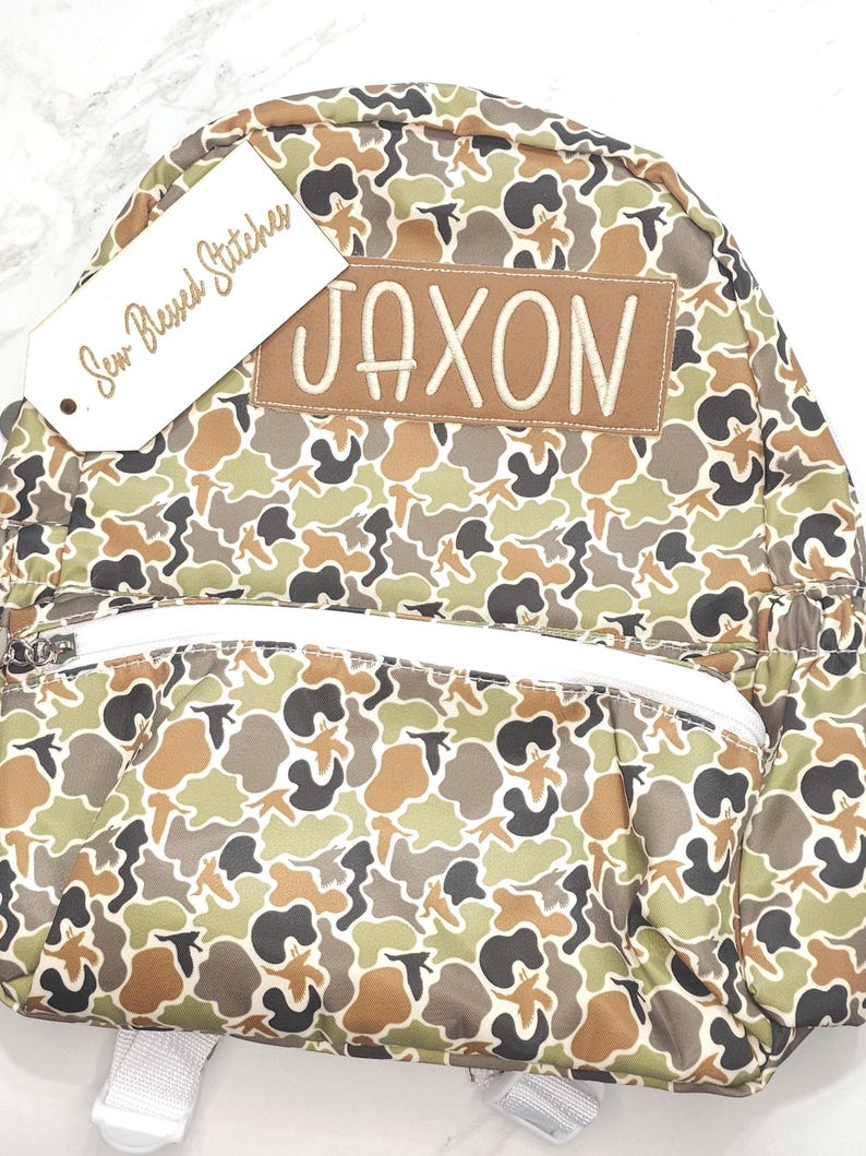 Puede incluir: Una mochila con estampado de camuflaje con el nombre "JAXON" bordado en un parche marr&oacute;n. La mochila tiene una cremallera y correas blancas, y una etiqueta que dice "Sew Blessed Stitches". El patr&oacute;n incluye tonos de verde, marr&oacute;n y negro.