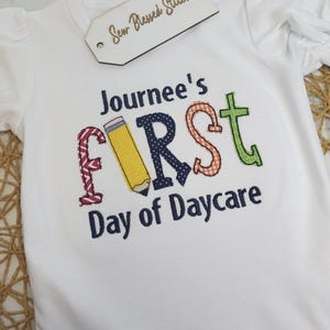 K&ouml;nnte beinhalten: Wei&szlig;er Strampler mit einem bunten gestickten Design, das "Journee's First Day of Daycare" mit einer Bleistiftgrafik sagt.
