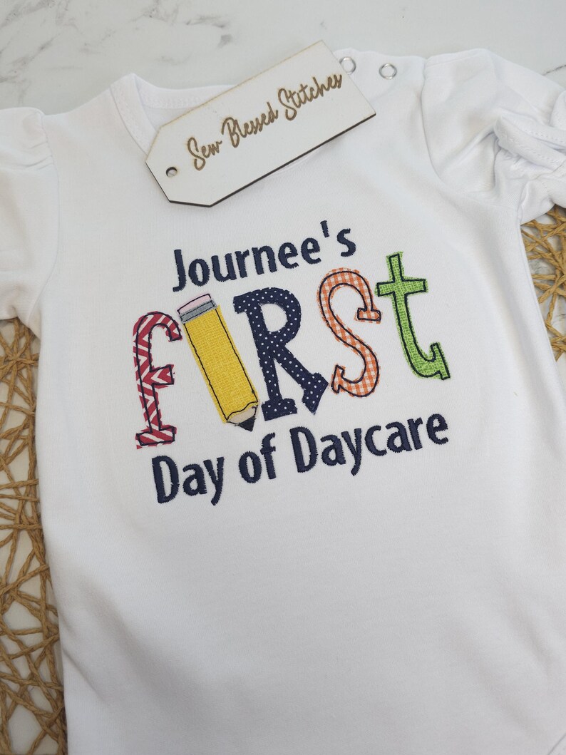 K&ouml;nnte beinhalten: Wei&szlig;er Strampler mit einem bunten gestickten Design, das "Journee's First Day of Daycare" mit einer Bleistiftgrafik sagt.