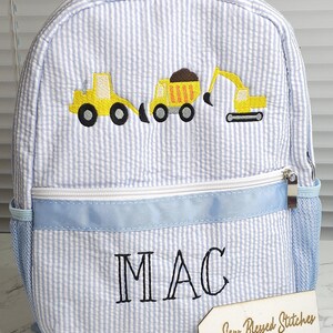 May include: Blue and white striped backpack with embroidered yellow construction vehicles. Features a blue ribbon accent and the name "MAC" embroidered in black. Includes a side mesh pocket and a zipper closure.