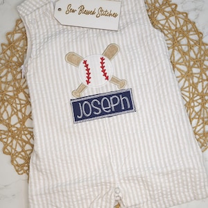 Peut inclure: Une barboteuse en seersucker à rayures beiges et blanches ornée d'une broderie représentant une balle de baseball et deux battes croisées au-dessus d'un écusson bleu marine avec le prénom JOSEPH brodé en blanc. Une étiquette Sew Blessed Stitches.