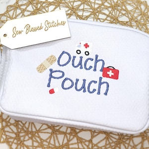 May include: A light blue "Ouch Pouch" with embroidered text and medical-themed graphics. The pouch features a bandage, pill, ambulance, and a red first aid kit. A tag reads "Sew Blessed Stitches".