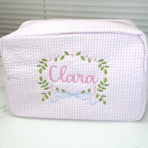 May include: A pink and white seersucker cosmetic bag with a floral wreath and the name "Clara" embroidered in pink. The bag has a zipper closure and a rectangular shape.