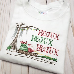 May include: A white short-sleeved t-shirt featuring an embroidered design of Santa Claus in a canoe pulled by an alligator. The design includes a cypress tree with Spanish moss, string lights, and the red and green text "HEAUX HEAUX HEAUX".