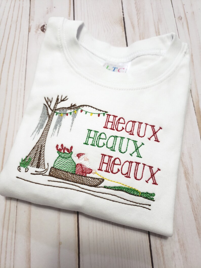 May include: A white short-sleeved t-shirt featuring an embroidered design of Santa Claus in a canoe pulled by an alligator. The design includes a cypress tree with Spanish moss, string lights, and the red and green text "HEAUX HEAUX HEAUX".