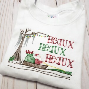 May include: A white t-shirt featuring an embroidered design of Santa Claus in a pirogue boat with a green sack, pulled by an alligator. A cypress tree with Spanish moss and string lights is visible. The text reads Heaux Heaux Heaux in red and green.
