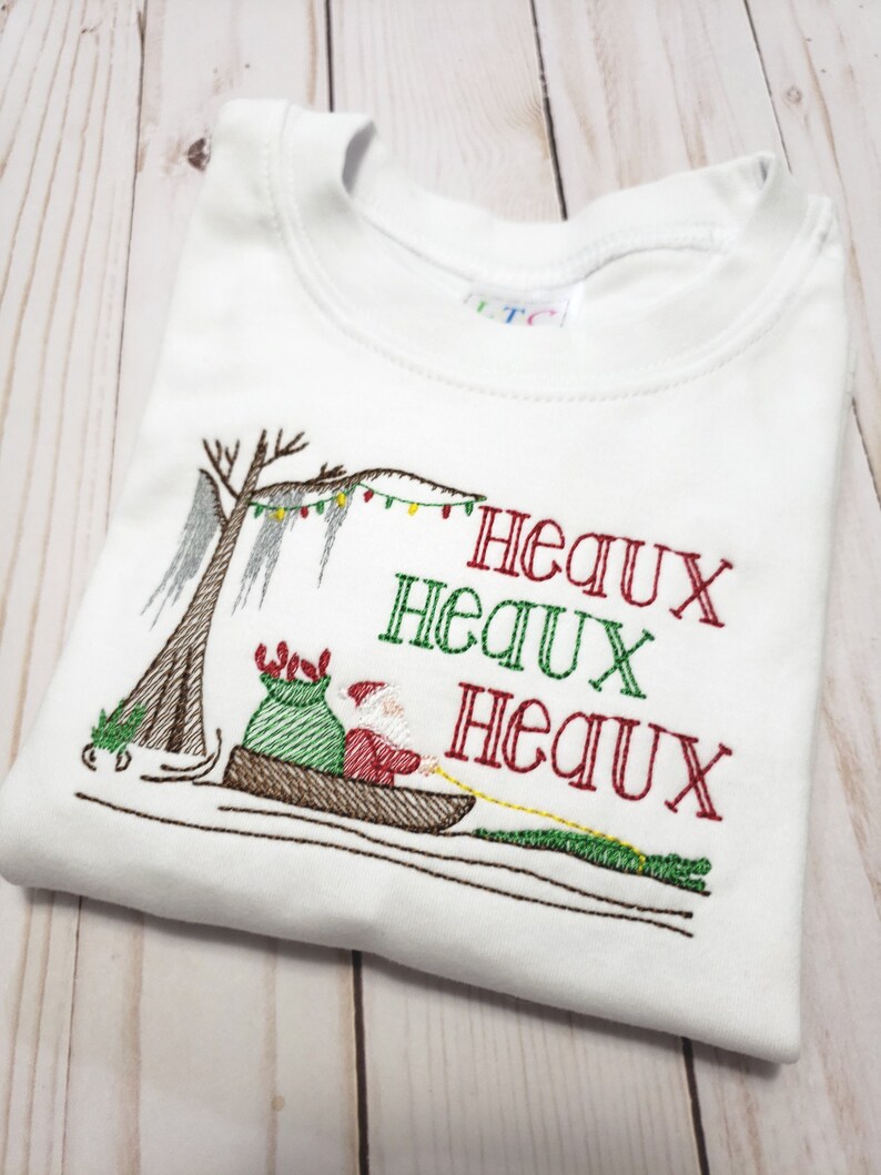 May include: A white t-shirt featuring an embroidered design of Santa Claus in a pirogue boat with a green sack, pulled by an alligator. A cypress tree with Spanish moss and string lights is visible. The text reads Heaux Heaux Heaux in red and green.