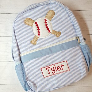 K&ouml;nnte beinhalten: Ein blauer und wei&szlig; gestreifter Rucksack mit einem Baseball- und Schl&auml;ger-Design. Der Rucksack hat eine blaue Netztasche an der Seite und ein rotes Namensschild mit der Aufschrift "Tyler".
