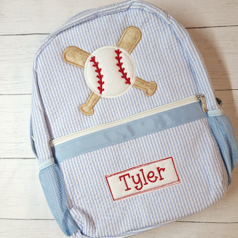 K&ouml;nnte beinhalten: Ein blauer und wei&szlig; gestreifter Rucksack mit einem Baseball- und Schl&auml;ger-Design. Der Rucksack hat eine blaue Netztasche an der Seite und ein rotes Namensschild mit der Aufschrift "Tyler".