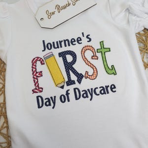 K&ouml;nnte beinhalten: Wei&szlig;er Strampler mit dem gestickten Text "Journee's First Day of Daycare" in bunten Buchstaben. Der Buchstabe "F" ist ein gelber Bleistift, der Buchstabe "I" ist rot-wei&szlig; kariert, der Buchstabe "R" ist blau mit wei&szlig;en Punkten, der Buchstabe "S" ist orange-wei&szlig; kariert und der Buchstabe "T" ist gr&uuml;n.