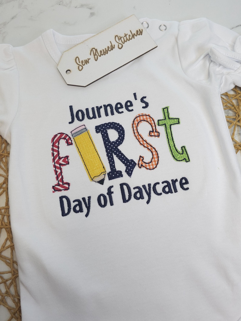 K&ouml;nnte beinhalten: Wei&szlig;er Strampler mit dem gestickten Text "Journee's First Day of Daycare" in bunten Buchstaben. Der Buchstabe "F" ist ein gelber Bleistift, der Buchstabe "I" ist rot-wei&szlig; kariert, der Buchstabe "R" ist blau mit wei&szlig;en Punkten, der Buchstabe "S" ist orange-wei&szlig; kariert und der Buchstabe "T" ist gr&uuml;n.