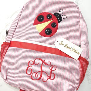 May include: A red and white pinstriped backpack featuring an embroidered ladybug applique on the top section and a red monogram with the letters E, J, and L on the front pocket. A white tag attached to the zipper reads Sew Blessed Stitches.